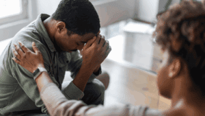 A person sits with head in hands, appearing distressed, while another offers comfort with a hand on their shoulder—a scene that could reflect the struggle behind determining a PTSD VA rating.