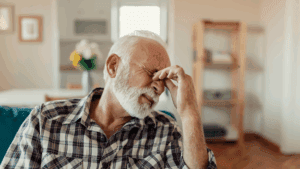 An older man with a white beard and plaid shirt sits on a sofa, pinching the bridge of his nose with his eyes closed, appearing to be in discomfort or stress, possibly struggling with migraines that affect VA disability ratings.