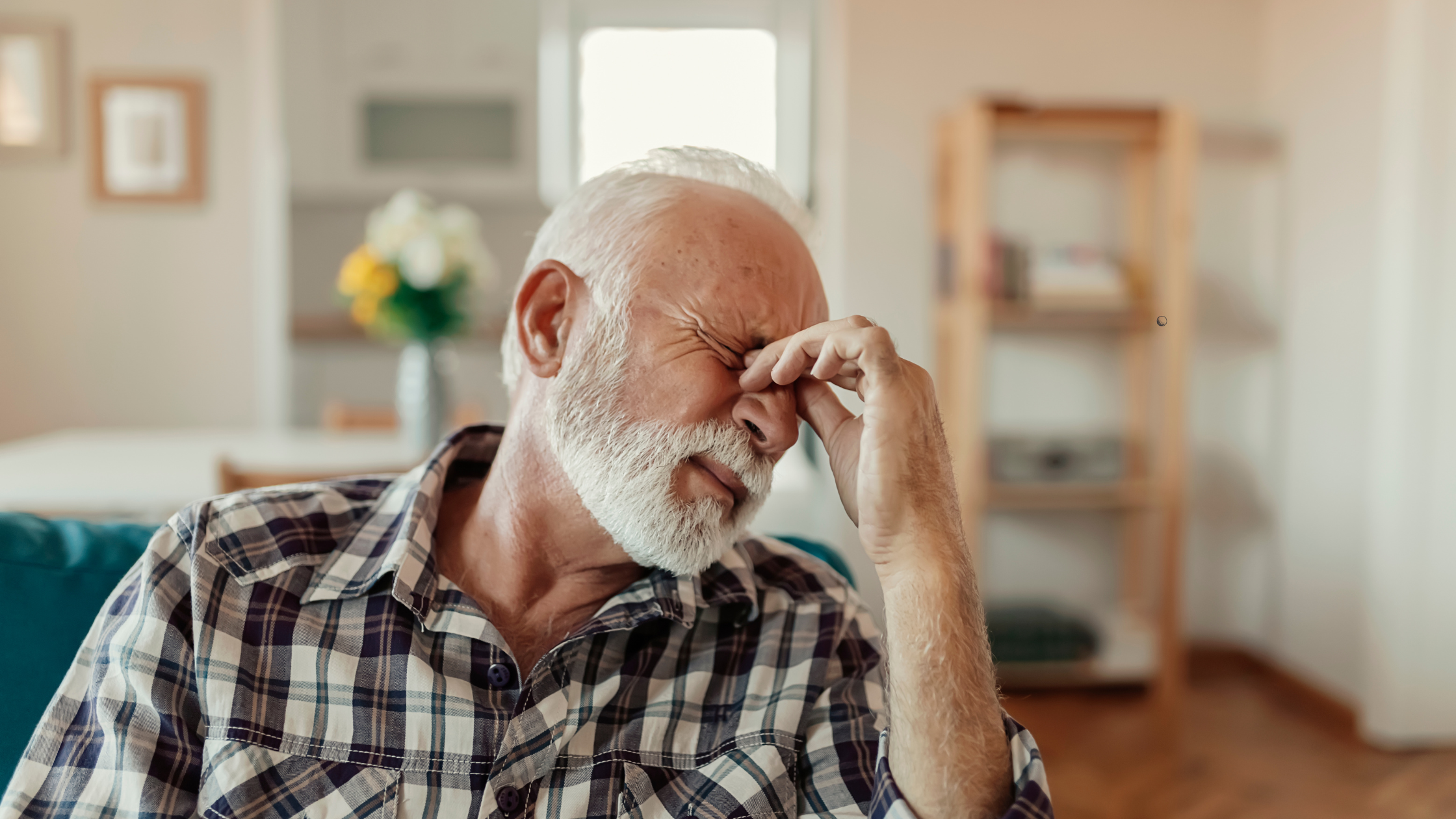 An older man with a white beard and plaid shirt sits on a sofa, pinching the bridge of his nose with his eyes closed, appearing to be in discomfort or stress, possibly struggling with migraines that affect VA disability ratings.