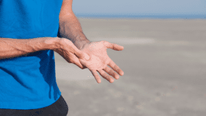 A close-up of a man's hands gesturing, with a blurred beach background. The man is wearing a blue shirt and in pain from carpal tunnel in his wrist.
