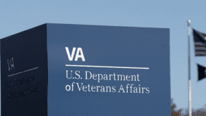 Signage for the U.S. Department of Veterans Affairs with the VA logo, set against a clear sky with American flags in the background, symbolizes unwavering support for those with 100% VA Disability.