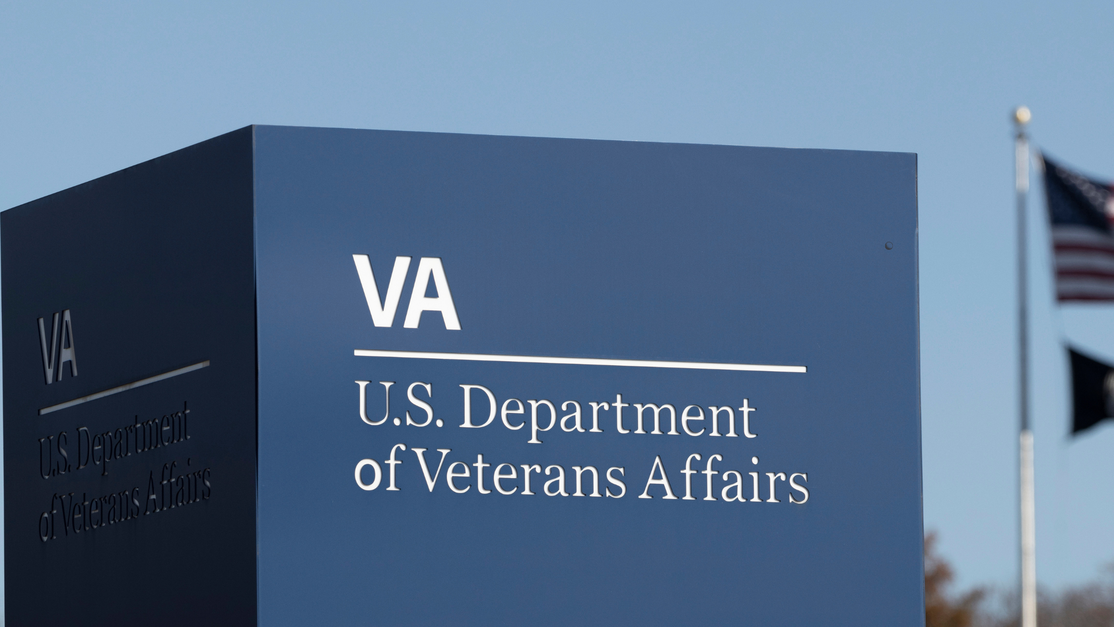 Signage for the U.S. Department of Veterans Affairs with the VA logo, set against a clear sky with American flags in the background, symbolizes unwavering support for those with 100% VA Disability.