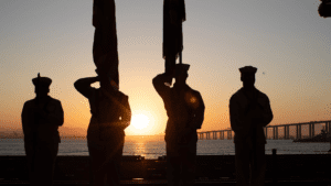 Silhouetted figures of 4 military service members, two saluting and two holding a flag, standing by a waterfront at sunset with a bridge in the background, reflecting on the new equity action plan.