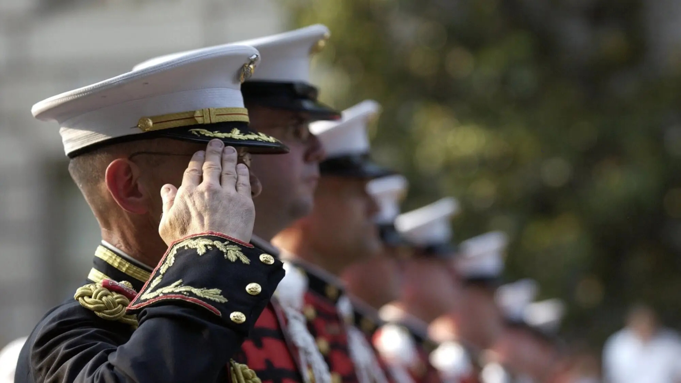 Uniformed military personnel saluting, with one in focus wearing a decorated uniform and white cap, as the 2025 COLA rates are announced