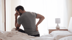 Person sitting on bed experiencing back pain, illustrating the impact of service-related conditions that may qualify for VA ratings for back pain. The individual is wearing a gray t-shirt and holding their lower back while appearing to be in discomfort.