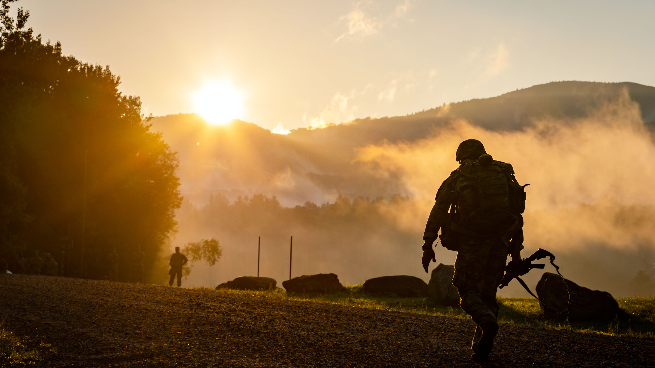 A soldier carrying a rifle walks along a gravel path at sunrise, contemplating the misty hills and trees in the background. The serene scene offers a momentary reprieve from thoughts of navigating his VA disability rating for depression.