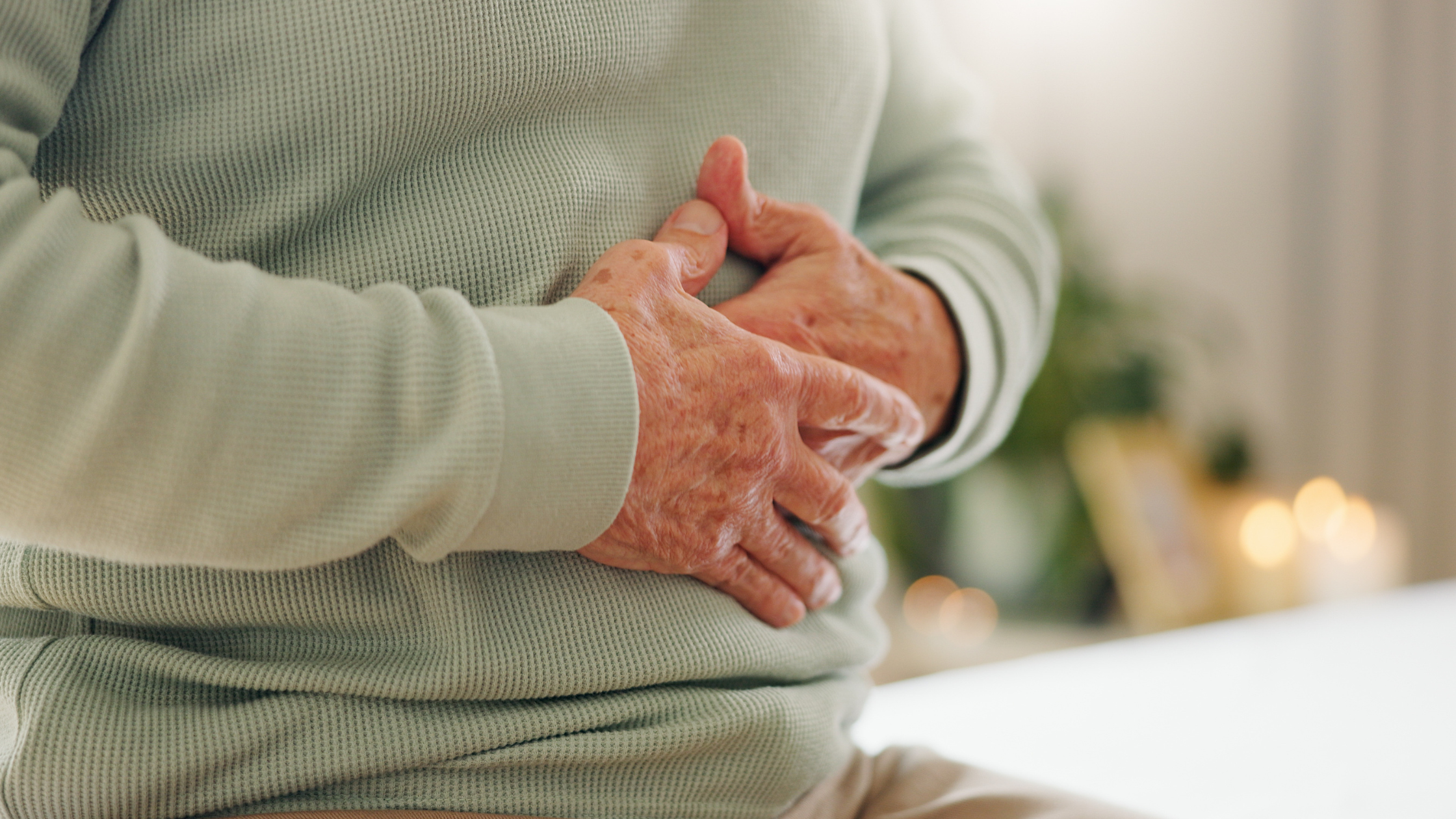 An elderly person in a light sweater clutches their stomach, seemingly experiencing discomfort or pain, perhaps wondering about the potential VA disability rating for IBS.