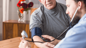 A doctor checks an older man's blood pressure with a sphygmomanometer in a room adorned with flowers, mindful of potential VA disability for hypertension concerns.