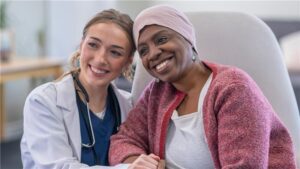 A doctor and a patient wearing a headscarf sit together, smiling and holding hands in a medical office while discussing VA Disability for Cancer.
