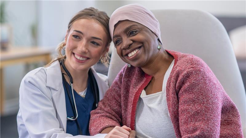 A doctor and a patient wearing a headscarf sit together, smiling and holding hands in a medical office while discussing VA Disability for Cancer.