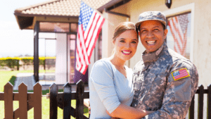 A smiling couple stands in front of a house with an American flag; one wears a military uniform, the other casual clothes, reflecting pride and support for veteran spouse benefits.